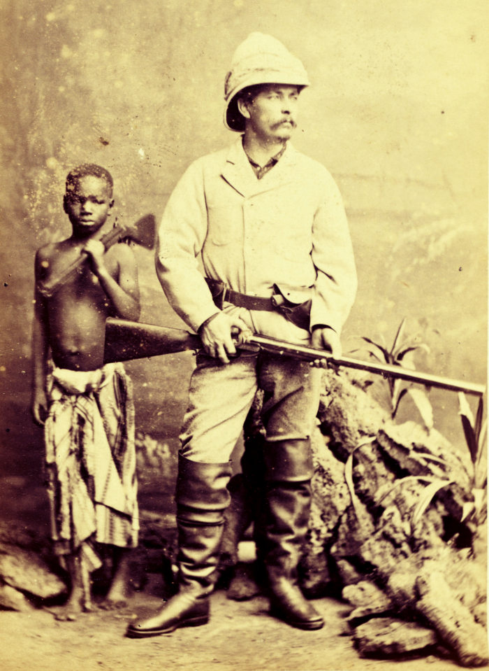 altes Foto von Henry Morton Stanley im Tropenanzug und mit Flinte, neben ihm ein afrikanischer Junge, ebenfalls mit Flinte